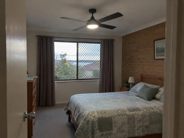 Bedroom with a ceiling fan above a bed, brick feature wall and window with curtains in Bonnells Bay
